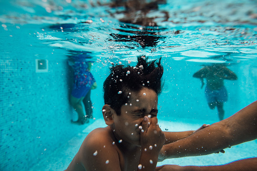 Close-up of boy holding breath underwater in a swimming pool. He is holding his nose and has his eyes closed. In the background, people are swimming. The boy is holding onto an arm as he is submerged.