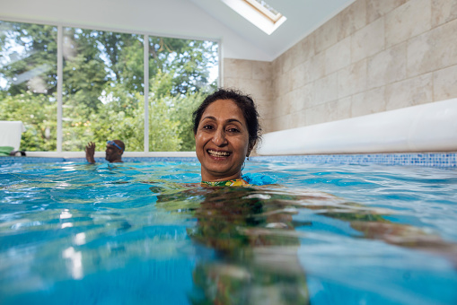 Wide shot of a mature woman smiling whilst swimming in a swimming pool. She is wearing modest swimwear as she is Muslim. Her body is fully submerged with a mature man in the background.