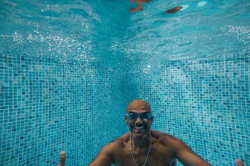 Close-up of a mature man playfully swimming underwater in a swimming pool. He is smiling towards the camera with his hands and arms pointing forward. He is wearing bright blue swimming goggles and a silver necklace.