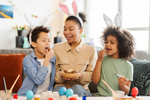 Portrait happy latin family eating chocolate eggs at home. Smiling mother and kids wearing bunny ears celebration Easter together. Holiday activity concept
