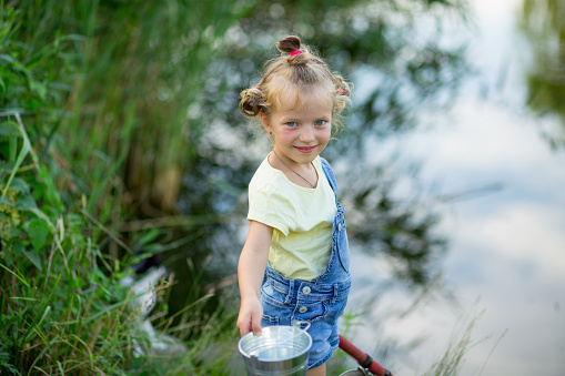 Three-year-old blond girl in denim shorts with straps catches fish on the river