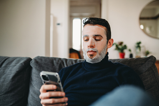 A young adult man is wearing a fabric face mask during his daily beauty routine. He's sitting on the sofa and he's typing on his smart phone.