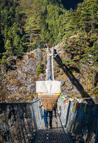 Sherpa porter carrying  expedition kit bags and equipment using a traditional basket across a wire bridge deep in the mountain wilderness of the Sagarmatha National Park, Himalayas, Nepal.