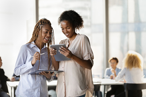 Happy young afro american females teammates enjoy solving business problem together using online and paper reports. Two millennial women employees feel glad to find perfect decision working together