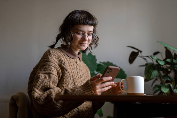 istock Pleased hipster woman drinking tea looking at smartphone screen smiling sitting at table at home. 2040848155