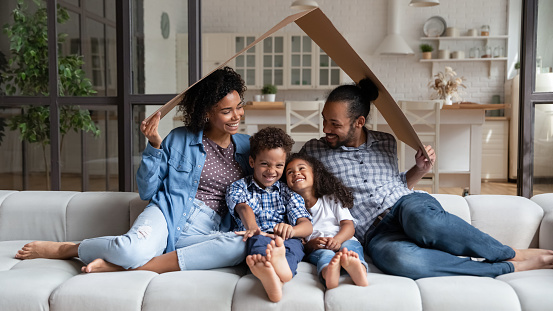 Happy affectionate young African American couple parents sitting under carton roof on comfortable couch with small adorable children son daughter, celebrating moving into own home, real estate concept