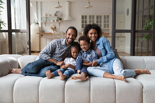 Full length portrait of smiling friendly African American family with cute little kids siblings relaxing on huge cozy couch in modern living room, enjoying carefree weekend time together at home.