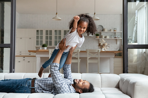 Cheerful little African American kid girl looking at camera playing super hero game, flying in arms of smiling affectionate caring daddy, having fun together on comfortable sofa on weekend at home.