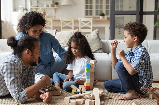 Happy young African American parents playing toys with adorable little preschool kid boy girl, enjoying playtime activity sitting on floor carpet in modern living room, family entertainment concept.