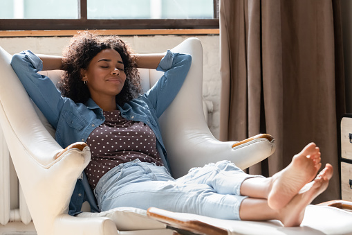 Full length millennial African American woman homeowner daydreaming in comfortable armchair with legs on footstool, breathing fresh air, sleeping resting, enjoying peaceful leisure time at home.