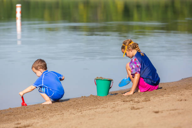 Cute young girl having fun on a sandy lake beach on warm and sunny summer day. Kid playing by the river. Summer activities for children.