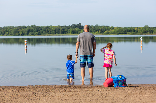 Rear view of a father and two young children standing in the shallow water at the beach on a late summer morning.