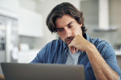 Serious, thinking and stressed man reading on laptop while sitting at home. Frustrated and upset man doing research on internet, checking email or waiting for online loan or job application answer