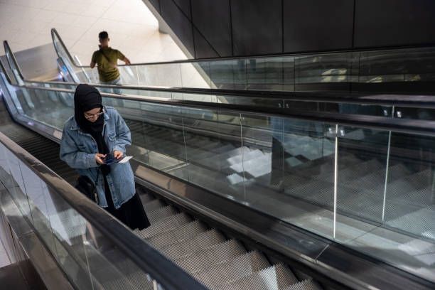 Young casual female using escalator in airport holding her passport and using her mobile phone during traveling