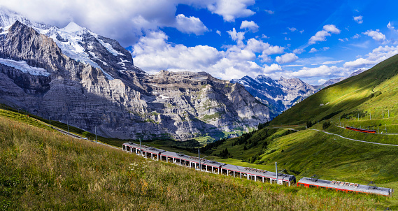 Kleine Scheidegg, Bernese Oberland, Switzerland - July 3, 2022:  The famous Red Jungfraubahn train that travels to and from Jungfraujoch and Kleine Scheidegg, passing through the Mountain Eiger and making it the highest railway line in Europe. Eiger and Jungfrau mountain peaks in the background.