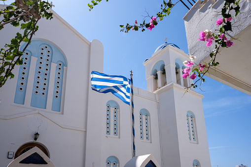 View of the greek flag waving in the air next to a bougainvillea and a whitewashed church in the background in Ios Greece