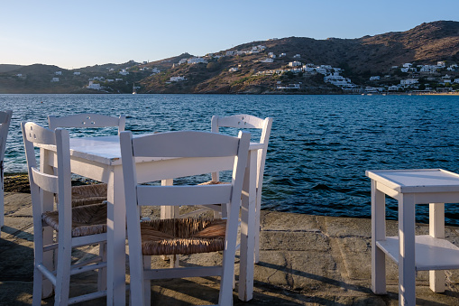 View of a restaurants white chairs, tables and the blue sea in the background in Ios Greece