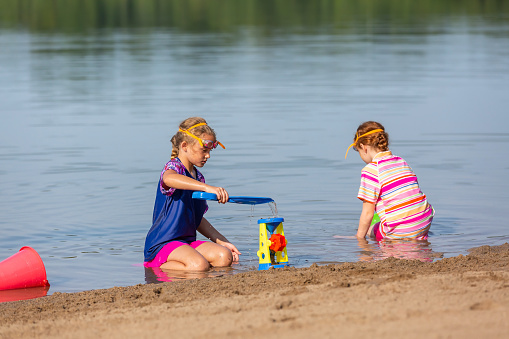Two young girls (sisters) sitting at the edge of the water playing with water and sand toys.