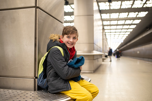 smiling 11 year old schoolboy in winter clothes sitting on a bench at a metro station, waiting for the train to arrive