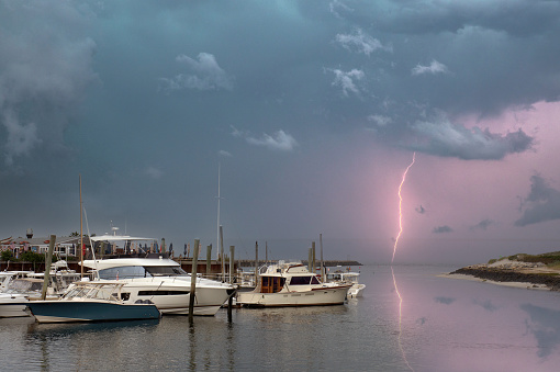 Dramatic Cape Cod coastal scene in East Dennis, Massachusetts. Bolt of lightning discharged from cloud in menacing dark gray sky above boats anchored in harbor.