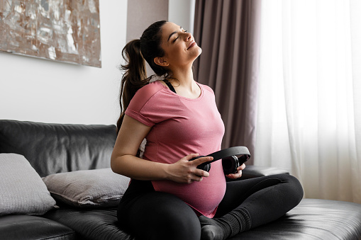 Nice adorable lovely sweet cheerful positive pregnant  mum sitting on sofa, couch, divan, keeping earphones on belly, classical music, white light interior room
