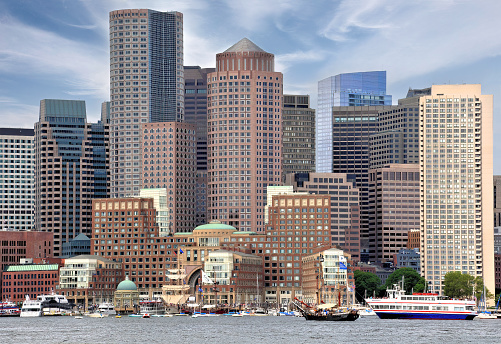 Colorful cityscape of Boston skyline and waterfront with tall modern buildings, boats anchored in harbor, and lovely blue sky overhead.