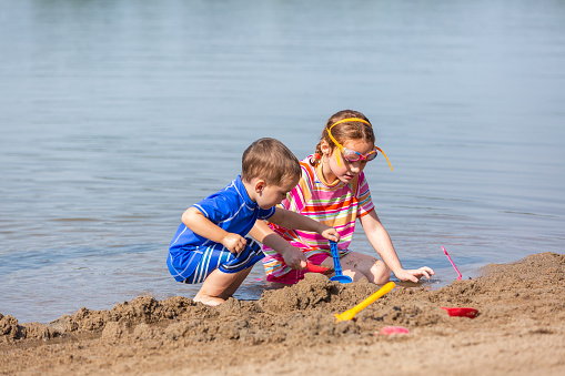 A young brother and sister at the edge of the water playing with water and sand toys.