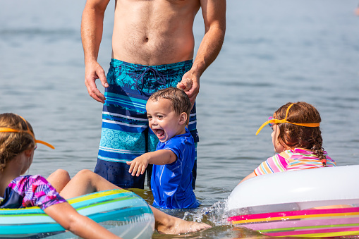 Close-up of a two year old boy holding Dad's hand as he stands in the water at the beach with his family.