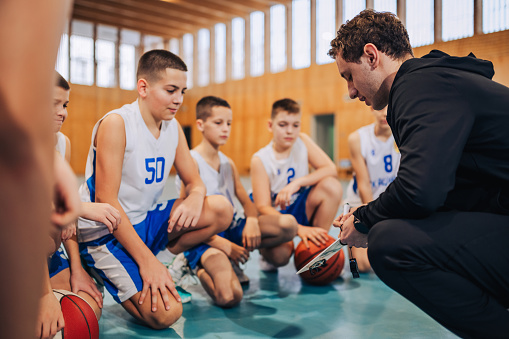 A junior basketball team is crouching on court with their young trainer and working on tactics and strategy. A basketball coach on court with his young team is working on game plan on training.