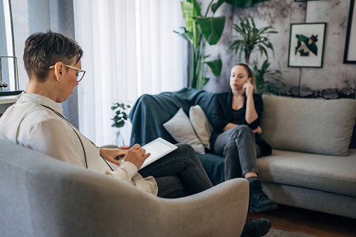 In a serene and inviting space, a therapist attentively listens to their client who is sharing their feelings. The room exudes tranquility with its soft lighting and comfortable furnishings, providing an ideal environment for healing and self-discovery.
