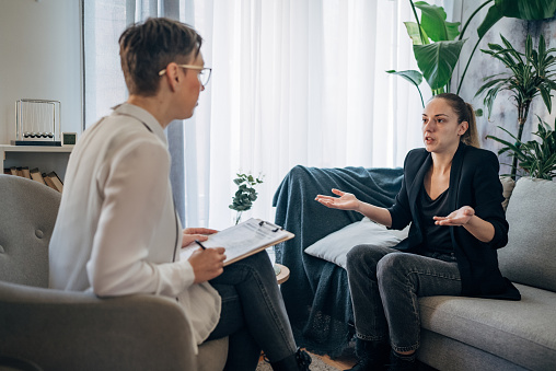 In a sunlit, cozy room, a therapist attentively listens to her client who is sharing personal experiences. The atmosphere is calm and supportive, fostering an environment of trust and healing.