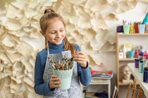 Smiling young girl holds cup of paintbrushes, surrounded by art supplies in creative classroom setting.