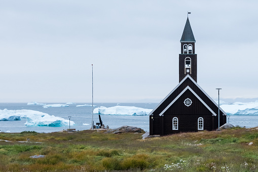 Illulisat, Greenland - Sep 7, 2023: Zion's Church is perched beside the waterfront in Illulisat.  Behind float icebergs recently liberated from the nearby, massive Icefjord.  Greenland.