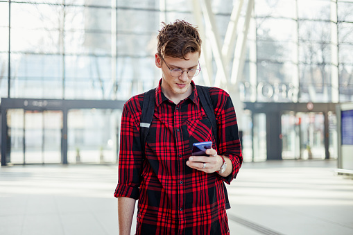 Portrait of a trendy young commuter standing at metro station with phone in his hands and typing messages. A passenger is standing at train station and using phone for telecommunications. Copy space.