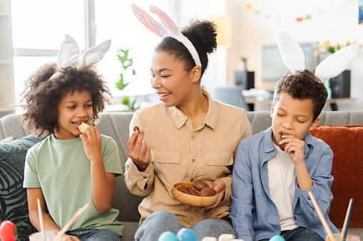 Portrait smiling African American family eating chocolate eggs at home. Happy mother and cute kids wearing bunny ears celebration Easter together
