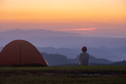 Traveller is sitting by the tent during overnight camping while looking at the beautiful scenic sunset over the mountain for outdoor adventure vacation travel concept