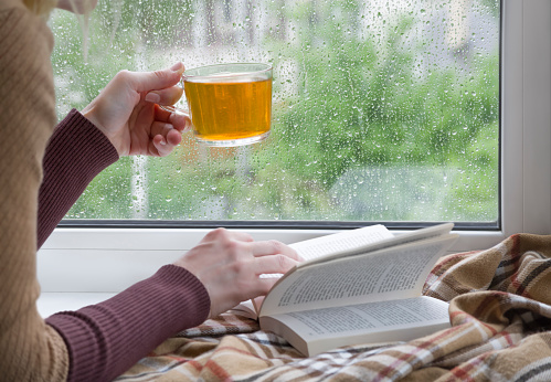 A woman with a cup of tea in her hand flips through a book lying on the windowsill, against the background of the window glass covered with raindrops