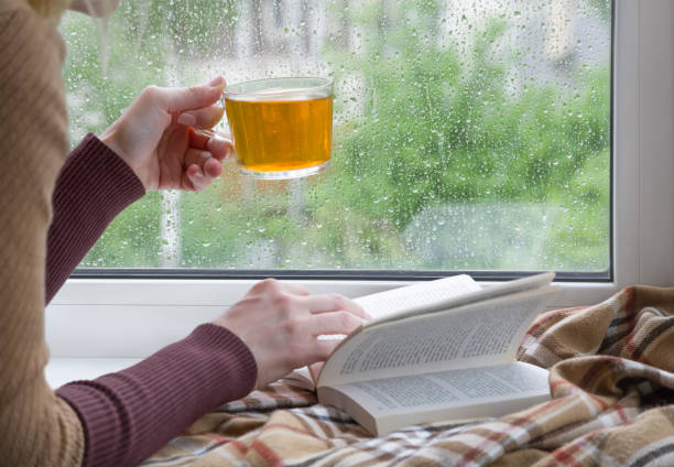 A woman's hand with a cup of tea. An open book and a soft blanket. The window glass is covered with raindrops. Springtime A woman with a cup of tea in her hand flips through a book lying on the windowsill, against the background of the window glass covered with raindrops woman hot shower stock pictures, royalty-free photos & images