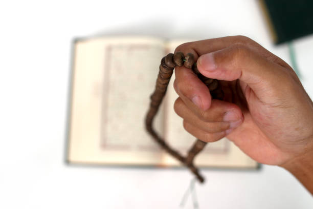 Muslim hand holding wooden prayer beads or Tasbih for dhikr during Ramadan, with a blurred Quran in the background, isolated on white