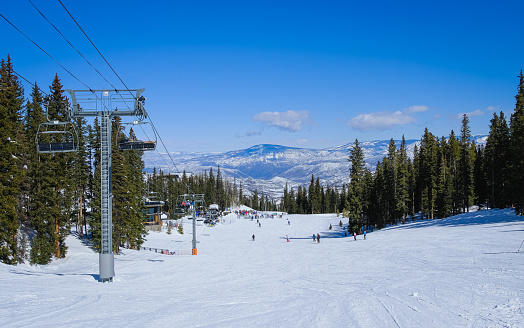 Beautiful view of Colorado ski resort on clear winter day; people skiing and snowboarding to base of chairlift; forest and mountains in background