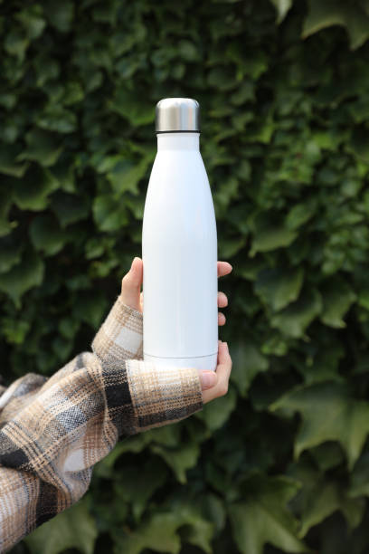 istock Hand holds white thermos bottle on green leaves wall background 2042051583