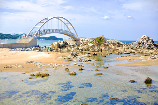 Yangyang County, South Korea - July 30th, 2019: An arched bridge extends to a rocky outcrop off the coast in Namae Port, with transparent waters in the foreground on a summer day.