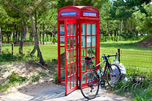 Yangyang County, South Korea - July 30th, 2019: The Jigyeong Park Certification Center, a distinctive red phone booth with a stamp for cyclists' passports, stands on sandy ground with green lawns and trees behind, a bike parked beside it.