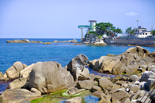 Yangyang County, South Korea - July 30th, 2019: The Namae Port Skywalk Observatory, a round structure with a glass-bottom platform extending 20 meters over the East Sea, stands against a rocky coastline and boulders in water on a summer day.