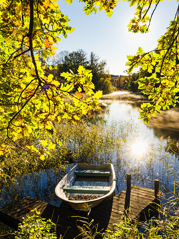 A tranquil scene unfolds along a river in Sweden, where a rowboat rests against a small wooden dock. Surrounded by the vibrant colors of autumn leaves and the gentle glitter of sunlight on the water, the setting exudes calm and natural beauty.