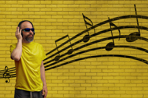 Bearded man with headphones listening to music standing in front of a yellow brick wall.