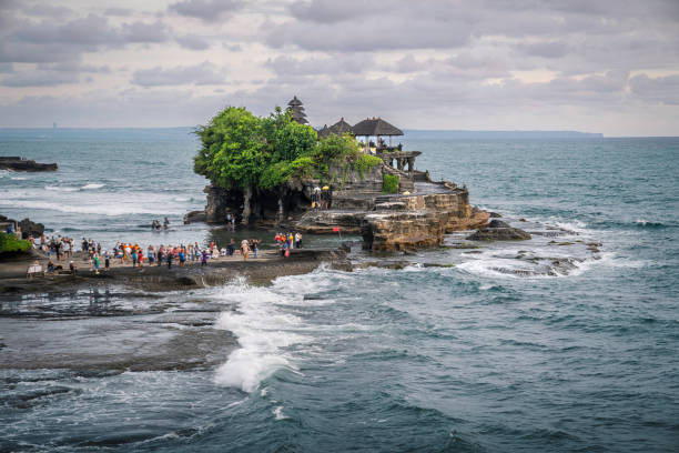 Tanah Lot temple on cliff in Indian ocean. Waves are breaking on a rock cliff off the coast of Bali. Hindu temple built on a rock in the sea. Lush tropical vegetation on top of a cliff.