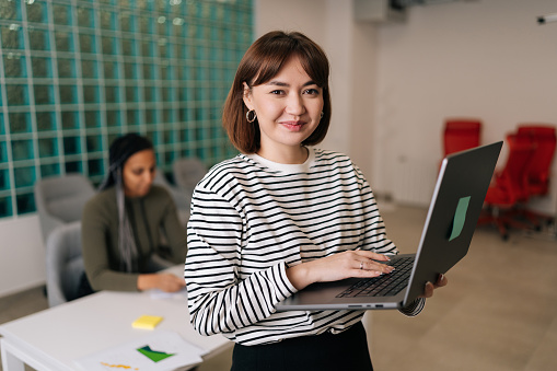 Portrait of friendly young businesswoman in casual clothes holding laptop in hand standing in office smiling looking at camera. Diverse business team discussing project sitting at desk on background