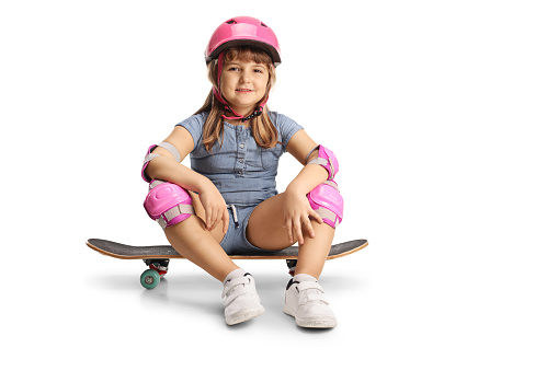 Full length profile shot of a little girl sitting on a skateboard, wearing elbow, knee pads and helmet isolated on white background