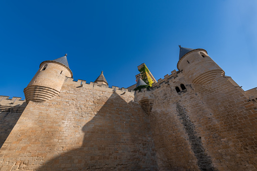 Beauvoir, France.  May 12, 2019.  Section of the castle of Mont St. Michel with flags blowing and people walking on the beach.  Sunny day with blue skies.